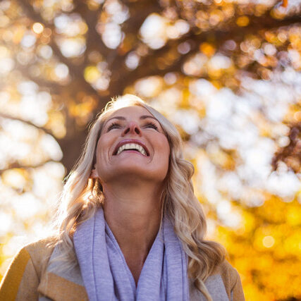 Smiling woman looking up against trees at park during autumn